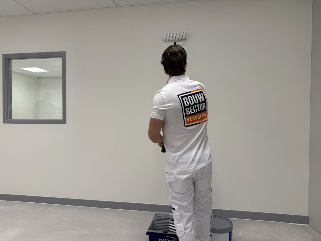 Schilder in witte werkbroek rolt een lichte wand naast een binnenraam, met de verfbak aan zijn voeten