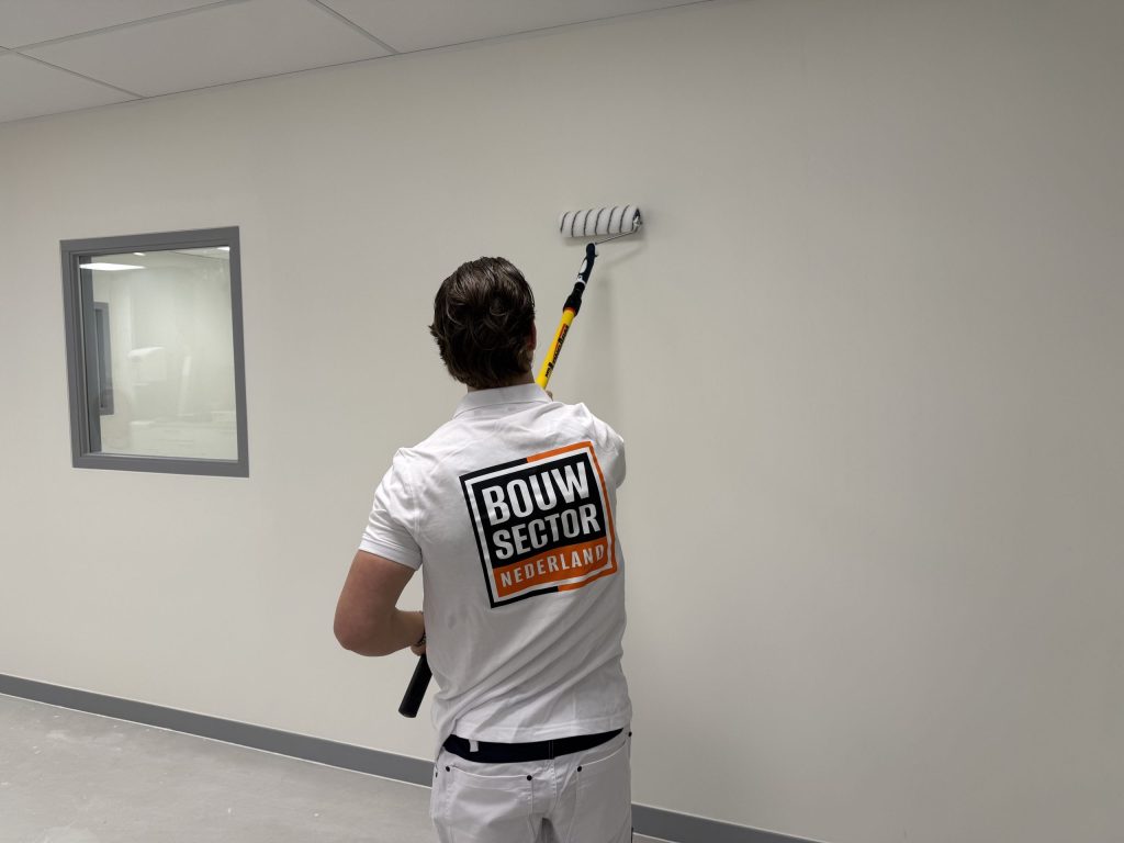 Schilder van Bouwsector Nederland zet met de verlengroller verf op een lichte wand naast een raam met grijs kozijn