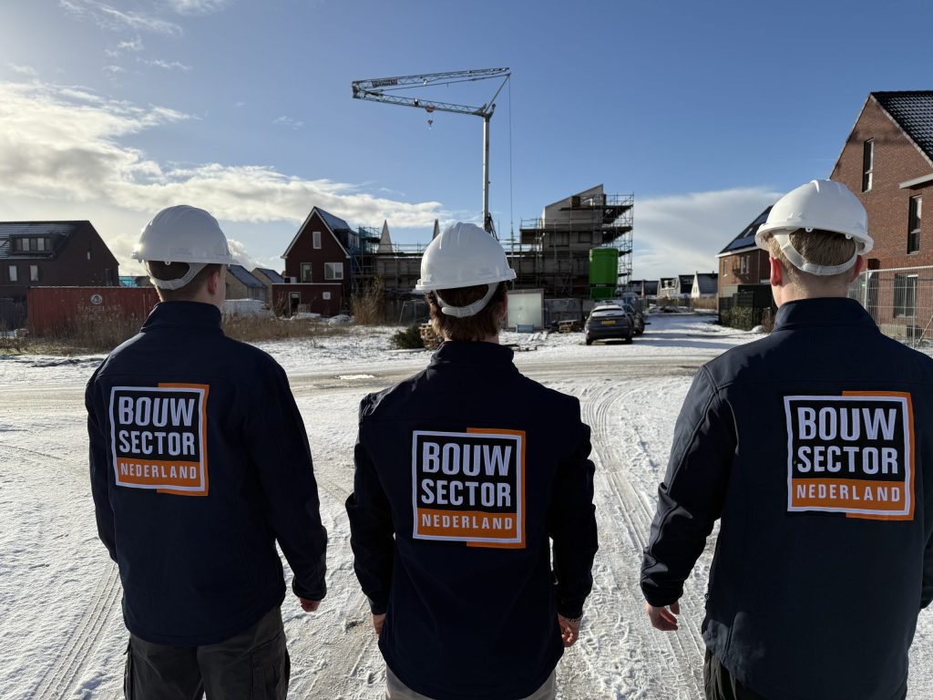 Drie mannen in jas van Bouwsector Nederland met bouwhelm kijken naar nieuwe huizen en een bouwkraan