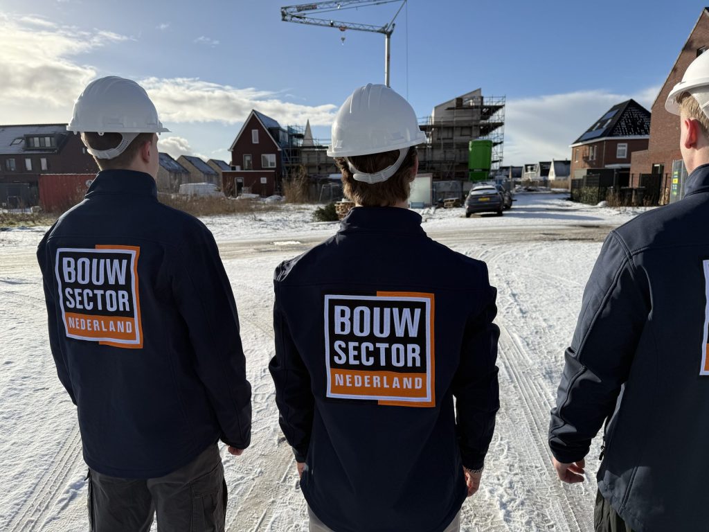 Drie vakmensen in bedrijfsjas kijken samen naar een woning in aanbouw op een winterse bouwplaats